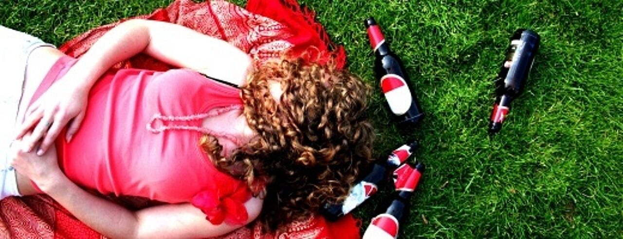 Blick von oben auf eine Frau, die auf dem Rücken im Gras liegt, mit langen Haaren, die ihr über dem Gesicht liegen, umringt von leeren Bierflaschen