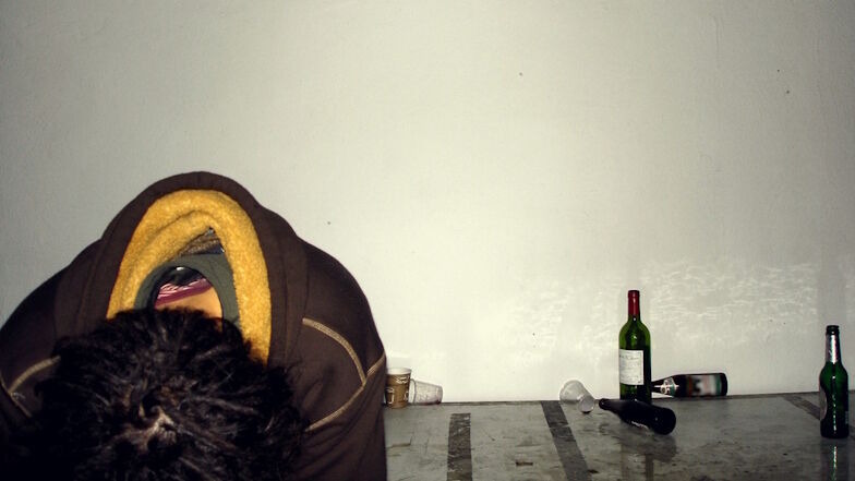 Leere Bier- und Weinflaschen in einem leeren Raum, im Vordergrund ein Mensch mit hängenden Kopf