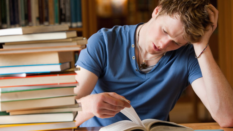 Männlicher Student sitzt in Bibliothek vor Bücherstapel, schaut müde auf ein Buch