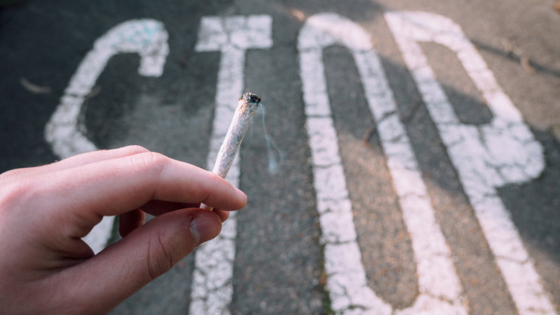 Hand hält brennende Zigarette vor auf der Straße aufgemaltem Wort "STOP"