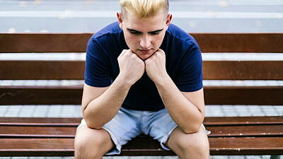 Junger Mann sitzt auf einer Bank, den Kopf auf beide Fäuste abgestützt und wirkt betrübt