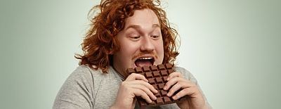 Übergewichtiger junger Mann beißt freudestrahlend in große Tafel Schokolade