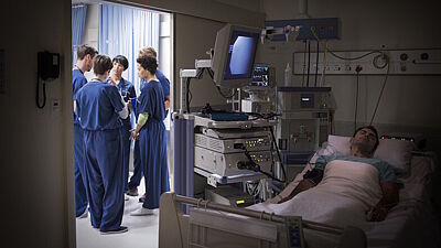 Mann liegt an Überwachungsgeräte angeschlossen und mit geschlossenen Augen im Krankenhausbett, während sich das behandelnde Team hinter geöffneter Tür unterhält.