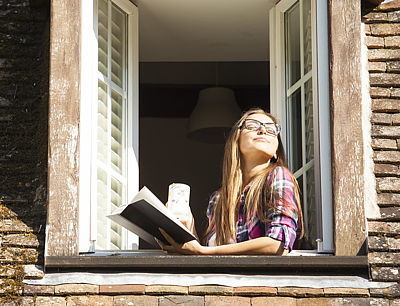 Frau lehnt sich aus dem Fenster und schaut mit Buch und Becher in der Hand in die Sonne