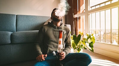 Mann sitzt auf dem Boden vor einem Sofa mit einer Bong vor sich und atmet Rauch aus