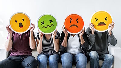 Vier Menschen sitzen aufgereiht nebeneinander und halten sich Emojis verschiedener Emotionen vor die Gesichter