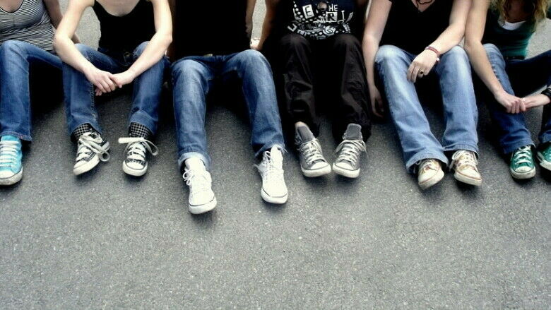 Sechs Jugendliche sitzen nebeneinander auf dem Asphalt; man sieht nur die Beine und den Oberkörper bis zu den Schultern