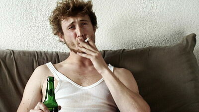 Junger Erwachsener mit Feinrippunterhemd auf Sofa und Bierflasche in der Hand zieht an Zigarette
