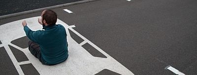 Mann sitzt auf einem auf der Straße aufgemaltem Auto und hält imaginäres Lenkrad in der Hand