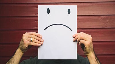 Mann hält Blatt Papier mit aufgemalten traurigen Smiley vor sein Gesicht