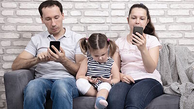 Bild: Vater, Mutter und kleines Kind auf Sofa, alle schauen auf ein Smartphone.
