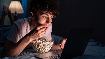 Junger Mann sitzt in dunklem Zimmer und isst Popcorn vor einem Laptop