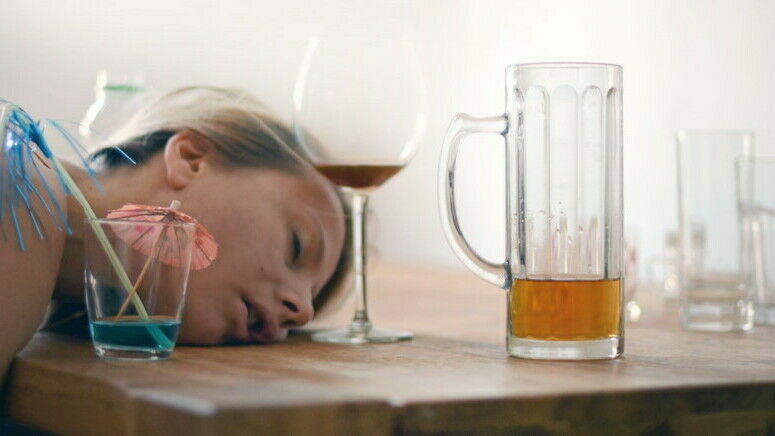 Jung Frau liegt nach einer Party anscheinend betrunken mit Kopf auf einem Tisch; darauf halbleere Gläser