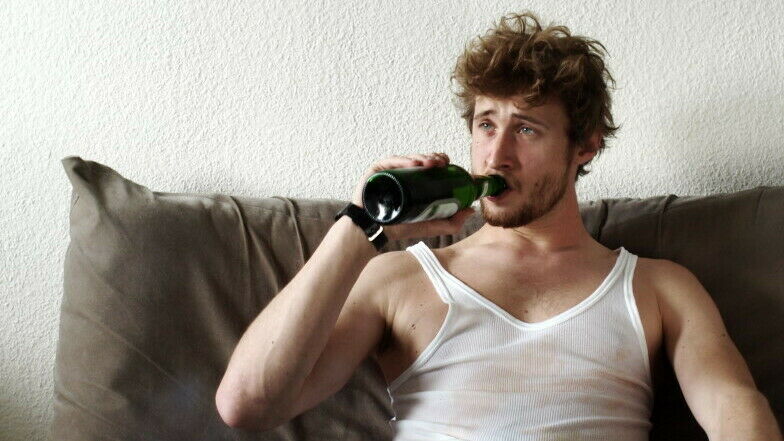Junger Mann sitzt mit schmuddeligem Unterhemd auf dem Sofa und trinkt aus einer Bierflasche