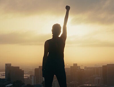Silhouette einer Frau in Siegerpose mit hochgerecktem Arm vor Sonnenauf- oder untergang und Scyline einer Stadt