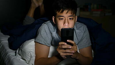 Junger Mann liegt im Dunkeln auf dem Bett und schaut auf sein Smartphone
