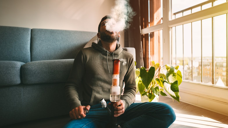 Mann sitzt auf dem Boden vor einem Sofa mit einer Bong vor sich und atmet Rauch aus