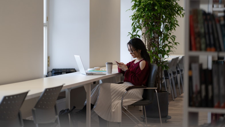 Studentin sitzt in einer Bibliothek mit dem Smartphone in der Hand