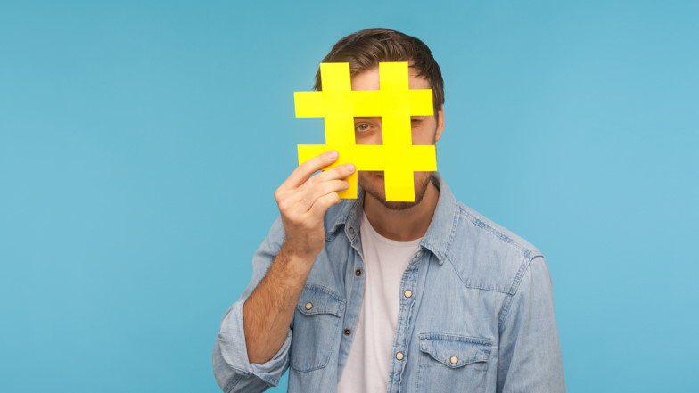 Junger Mann hält ein ausgeschnittenes Hashtag-Zeichen hoch und guckt mit einem Auge durch