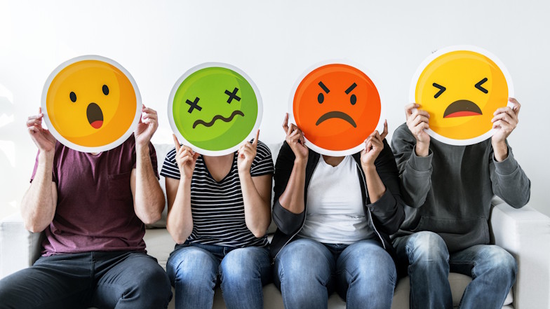 Vier Menschen sitzen aufgereiht nebeneinander und halten sich Emojis verschiedener Emotionen vor die Gesichter