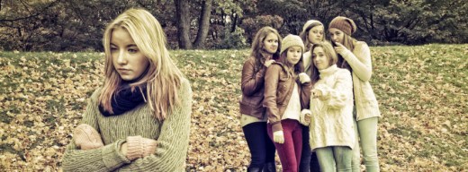Jugendliches Mädchen im Vordergrund, dahinter eine Gruppe Mädchen, die tuscheln und mit dem Finger auf das andere Mädchen zeigen