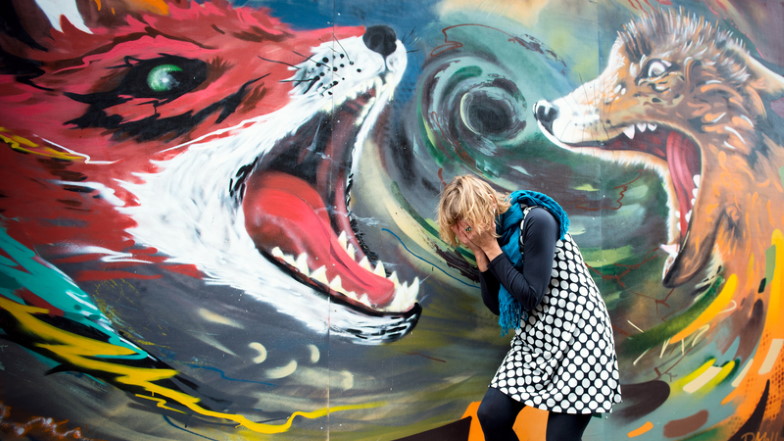 Frau steht gekrümt vor einer Wand mit großflächig gemalten Füchsen, die ihre Maul aufreißen und verbirgt ihr Gesicht in ihren Händen