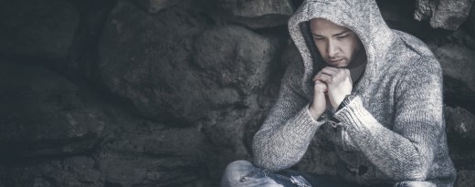 Junger Mann mit Kapuzenstrickpulli hokst vor einer Steinwand und schaut nach unten