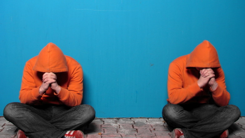 Zwei Männer in identischen orangen Hoodys sitzen im Schneidersitz mit gesenktem Kopf vor einer blauen Wand