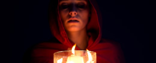 Frau steht vor dunklem Hintergrund mit roter Kapuze vor einer Kerze