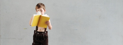 Kleiner Junge mit Lederhose vor Betonwand liest in großem Buch mit gelben Umschlag