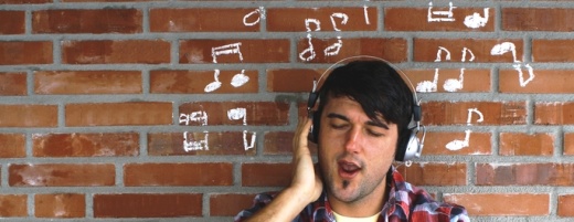 Mann hört Musik über Kopfhöhrer und steht vor Backsteinwand mit aufgemalten Noten