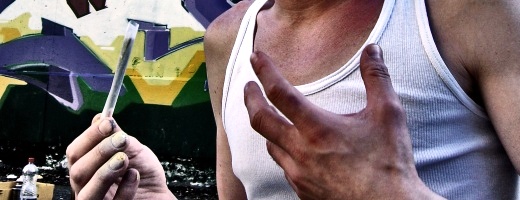 Oberkörper eines jungen Mannes im Unterhemd, der mit der linken Hand gestikuliert, in der rechten Hand einen Joint hält.