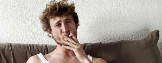 Junger Erwachsener mit Feinrippunterhemd auf Sofa zieht an Zigarette