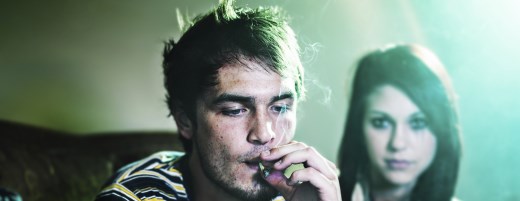Junger Mann raucht Joint, im Hintergrund schaut eine Frau zu