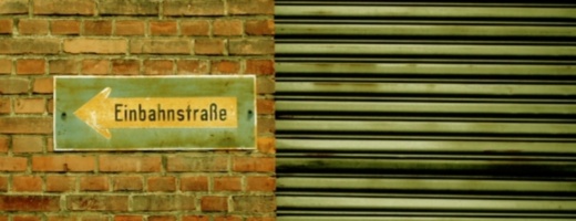 Vergilbtes Einbahnstraßenschild an Backsteinmauer, daneben heruntergelassener Rollladen