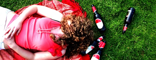 Blick von oben auf eine Frau, die auf dem Rücken im Gras liegt, mit langen Haaren, die ihr über dem Gesicht liegen, umringt von leeren Bierflaschen Blick von oben auf eine Frau, die auf dem Rücken im Gras liegt, mit langen Haaren, die ihr über dem Gesicht liegen, umringt von leeren Bierflaschen
