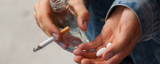 Nahaufnahme einer Person, die eine Zigarette, eine Schnapsflasche und Tabletten in den Händen hält