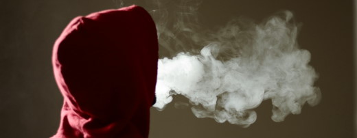 Person mit Kapuze über dem Kopf stößt Dampf einer E-Zigarette aus